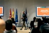 A person presenting at a conference with flags of Spain, the European Union, and another in the background.