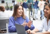 Two women are engaging in a conversation at an outdoor café with a laptop on the table.
