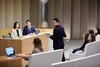 A formal meeting takes place with several individuals seated and a presenter standing in front.