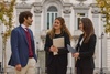 Three young professionals are having a conversation outdoors, surrounded by trees and a historic building.