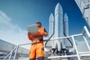 A technician in an orange uniform is using a laptop while standing near a large rocket.