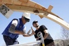 Two men constructing a wooden structure, one wearing a blue vest and a hard hat, the other in black, with QR codes visible on the structure.
