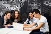 A group of four young adults is engaged in a collaborative discussion around a laptop in a modern office setting.