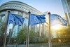 Three European Union flags flutter in front of a modern glass building on a sunny day.