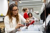 A woman is working on an electronic project at a modern workspace with colleagues in the background.