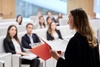A woman is presenting to an audience in a seminar hall, holding a red folder and addressing attendees who are seated and listening attentively.
