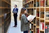 A woman reading a book in a library while a man stands nearby with a laptop.