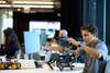 A young man assembles a robotic device at a workspace with various technology items in the background.