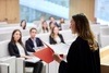 A woman in formal attire speaks to an audience in a modern setting.