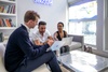 Three people are engaged in a discussion while working on laptops in a modern office setting.