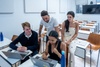 A group of four young adults collaborate around a laptop in a modern classroom setting.