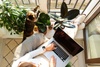 A person working on a laptop while sitting on a balcony surrounded by plants and a cat.