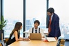 Three professionals are engaged in a discussion around a laptop in a modern office setting.