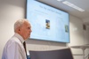An elderly man in a formal shirt sits in front of a presentation screen.