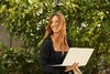 A young woman smiles while holding a laptop, standing outdoors surrounded by green foliage.