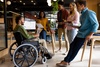 A group of five young professionals are collaborating in a modern office environment, with one person in a wheelchair actively participating in the discussion.