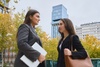 Two women are having a conversation while standing in an urban area with buildings in the background.