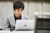 A young man in a suit sits at a table working on a laptop.