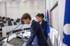 Two individuals are standing at a podium in a conference room, with flags in the background.