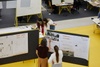 Three people are discussing over architectural drawings displayed on panels in a busy workshop environment.