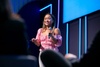 A woman in a pink blouse is speaking at a conference while holding a microphone.