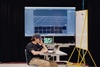 A young man is sitting at a table working on a laptop with a large screen displaying a digital interface in the background.