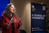 A woman in a red blazer is speaking passionately at an event related to humanities.
