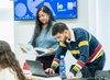 A student assists another student with a laptop in a university classroom.
