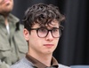 A young man with glasses and curly hair looks thoughtfully to the side.