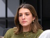 A young woman with brown hair wearing a green top sitting indoors, looking thoughtful.