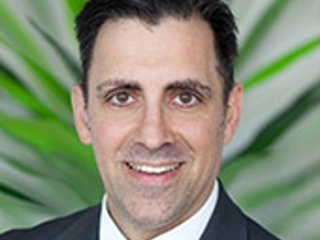 A professional portrait of a smiling man wearing a suit, with a tropical plant in the background.