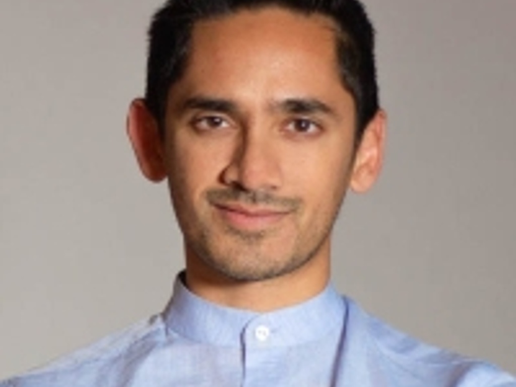 A portrait of a man with a light stubble, wearing a light blue buttoned shirt, smiling gently at the camera.