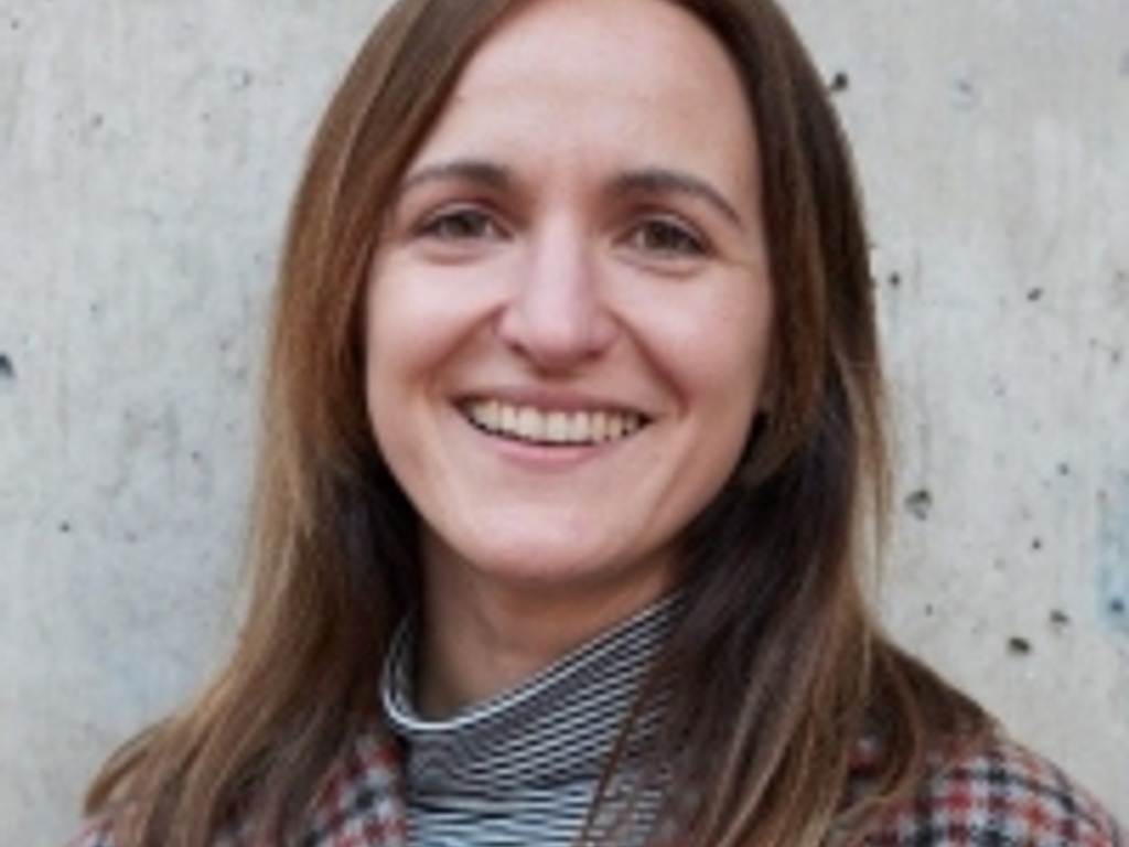 A smiling woman with brown hair wearing a checkered jacket and a striped shirt, standing against a concrete wall.