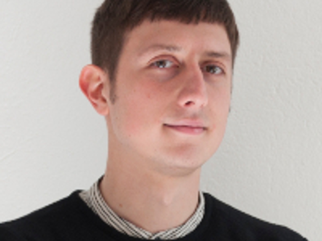 A portrait of a young man with a subtle smile, wearing a black sweater over a collared shirt, against a white background.