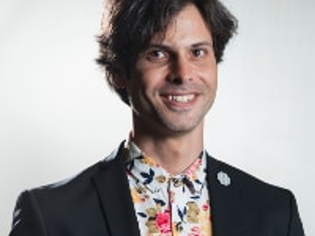 A man in a dark blazer and floral shirt smiles at the camera.