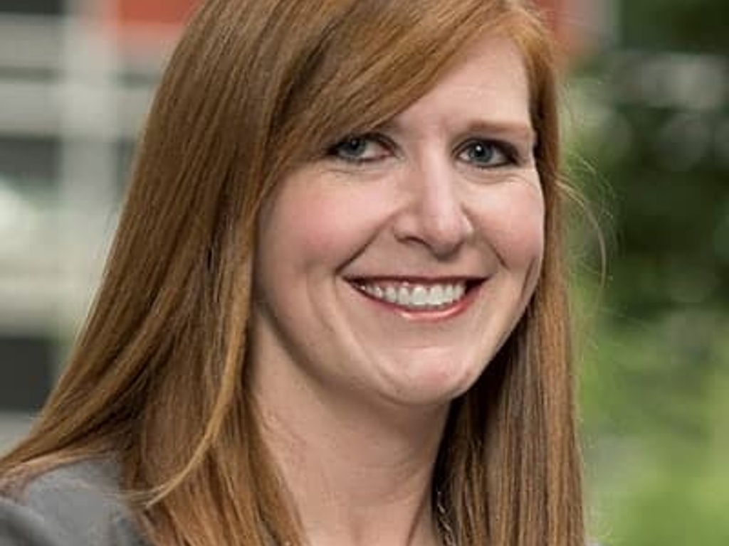 A portrait of a smiling woman with long red hair and a grey blazer.