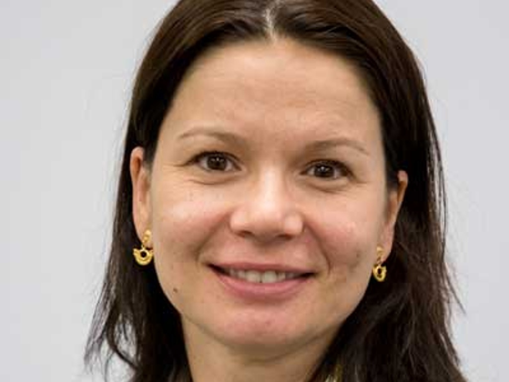 A portrait of a smiling woman with dark hair wearing earrings and a light-colored jacket.