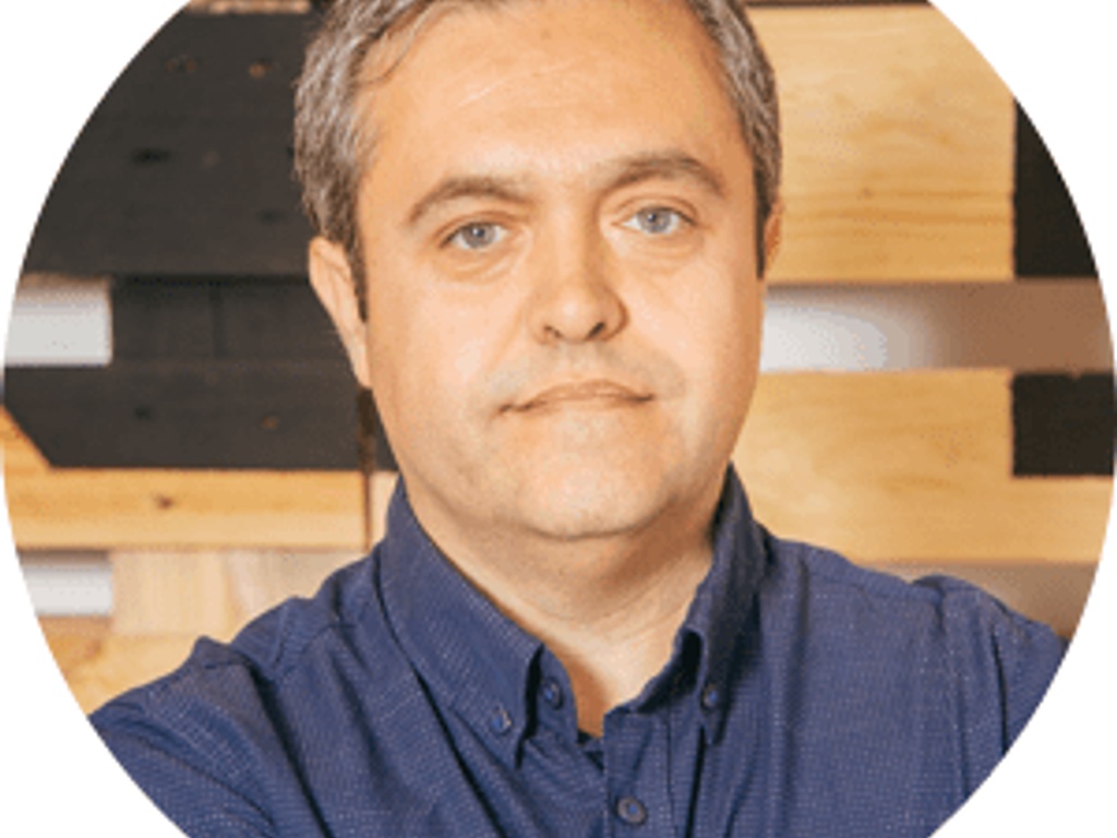 A middle-aged man in a blue shirt, looking directly at the camera with a subtle smile, set against a wooden background.