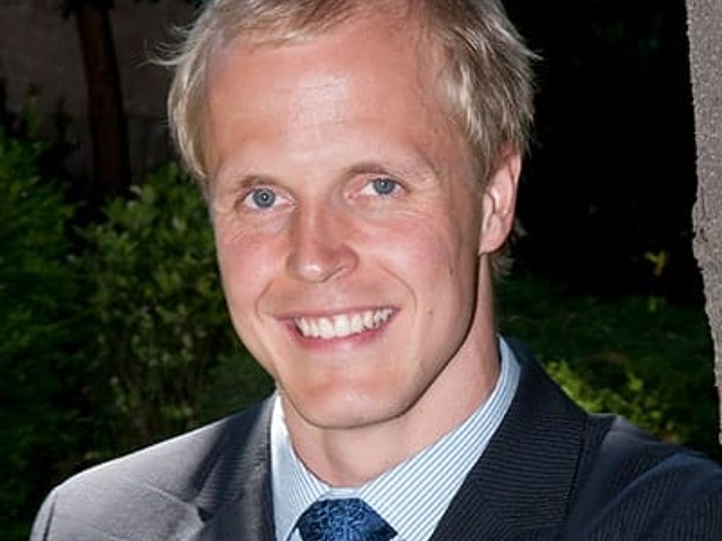 A smiling blond man in a suit and blue tie standing outdoors.