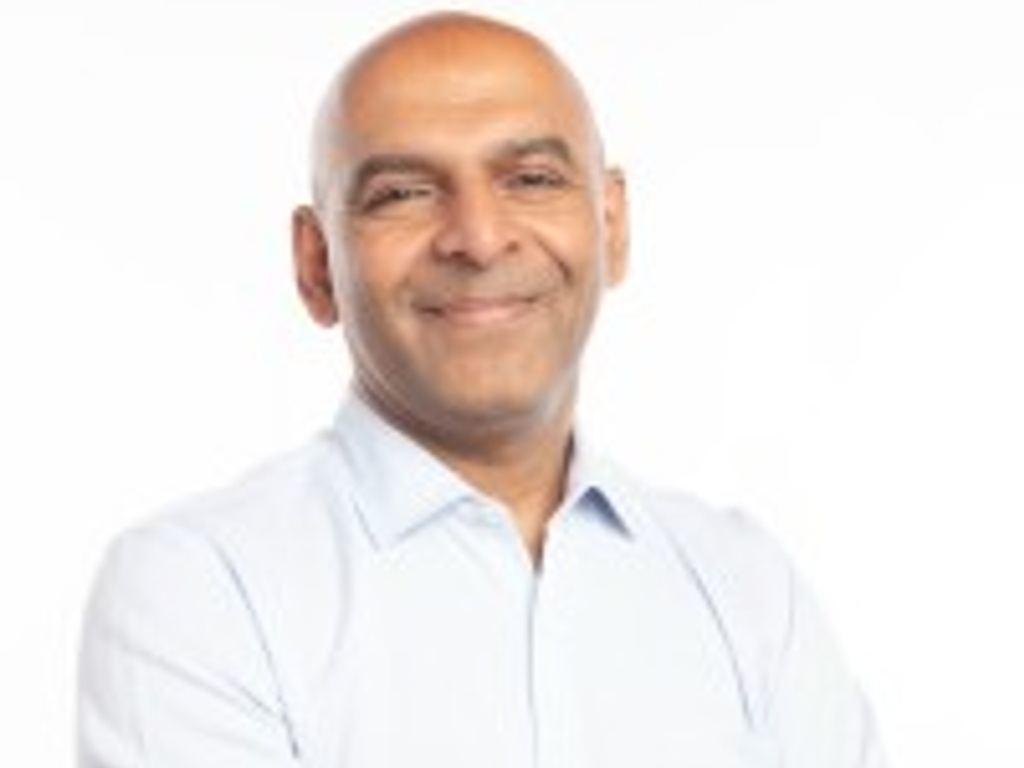 A smiling bald man in a light blue shirt with arms crossed standing against a white background.