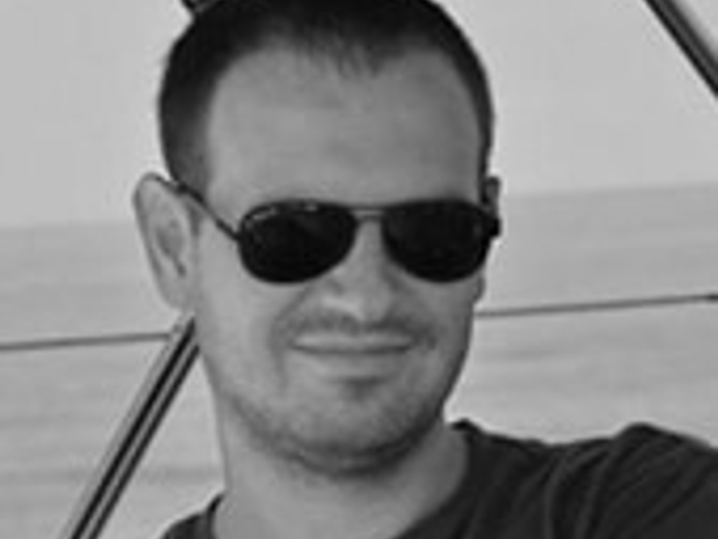 A man wearing sunglasses smiles slightly at the camera on a sunny day with a blurred background of sailboat rigging.