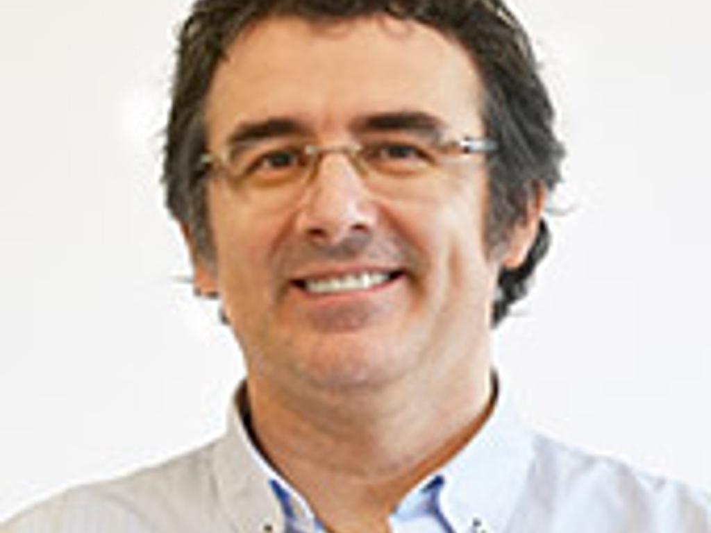 A portrait of a smiling man wearing glasses and a light blue collared shirt, standing against a white background.