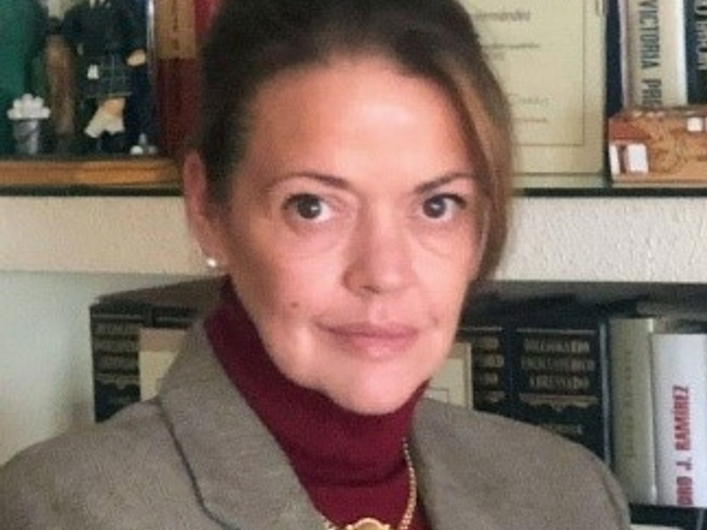 A professional woman with a calm expression wearing a blazer, standing in an office-like environment with books and a figurine in the background.