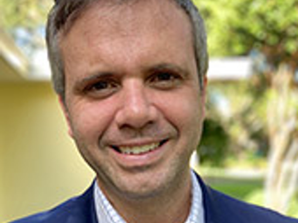 A smiling man wearing a blue blazer and checked shirt stands outdoors with greenery in the background.