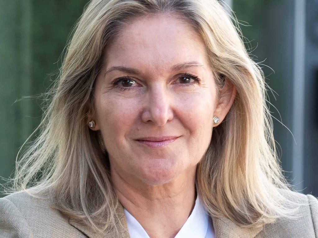 A portrait of a smiling middle-aged woman with blonde hair, wearing a beige blazer and a white shirt, standing outdoors.