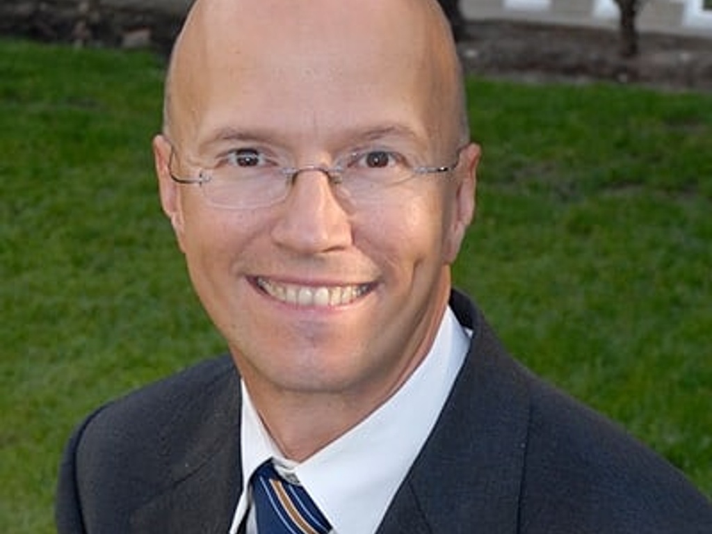 Portrait of a smiling bald man wearing glasses, a dark suit, and a striped tie in front of a blurred greenery background.