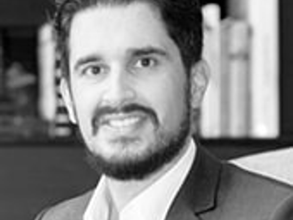 A professional black and white portrait of a smiling man with a beard, dressed in a suit and tie, standing in front of bookshelves.