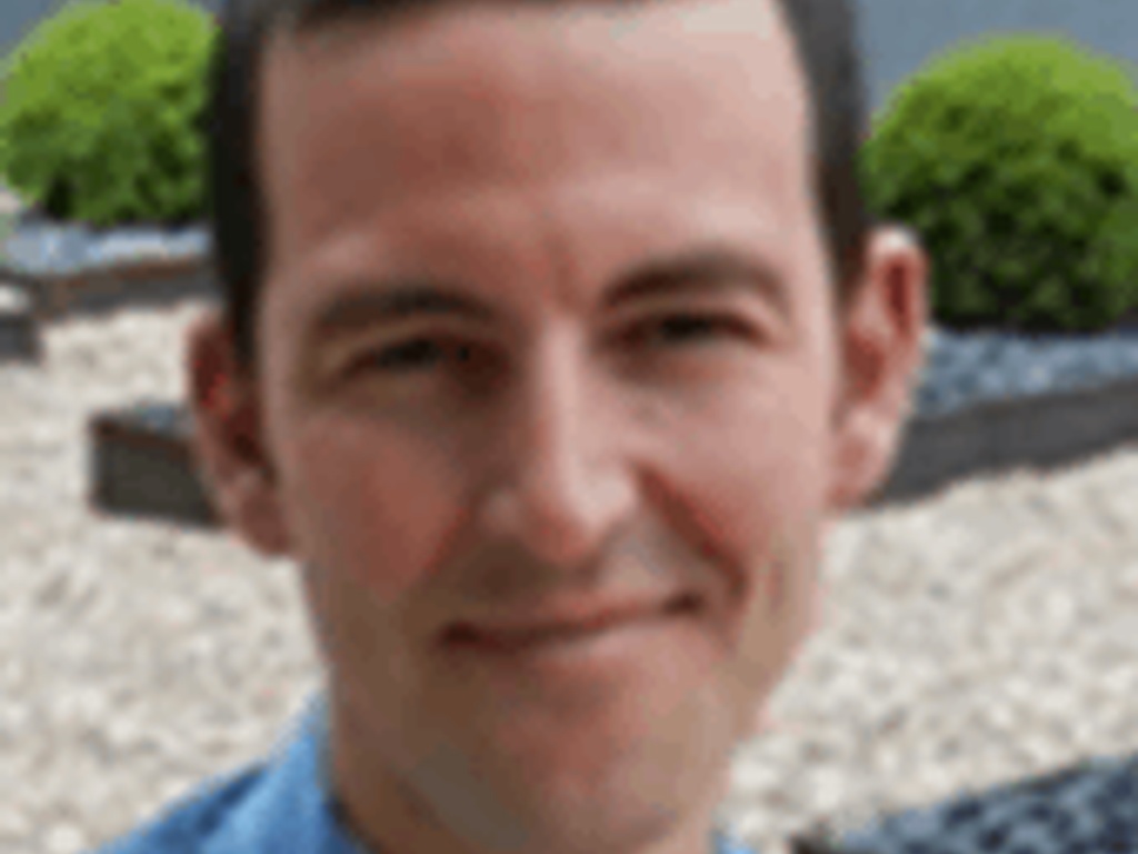 A smiling man in a blue sweater standing outdoors with plants in the background.