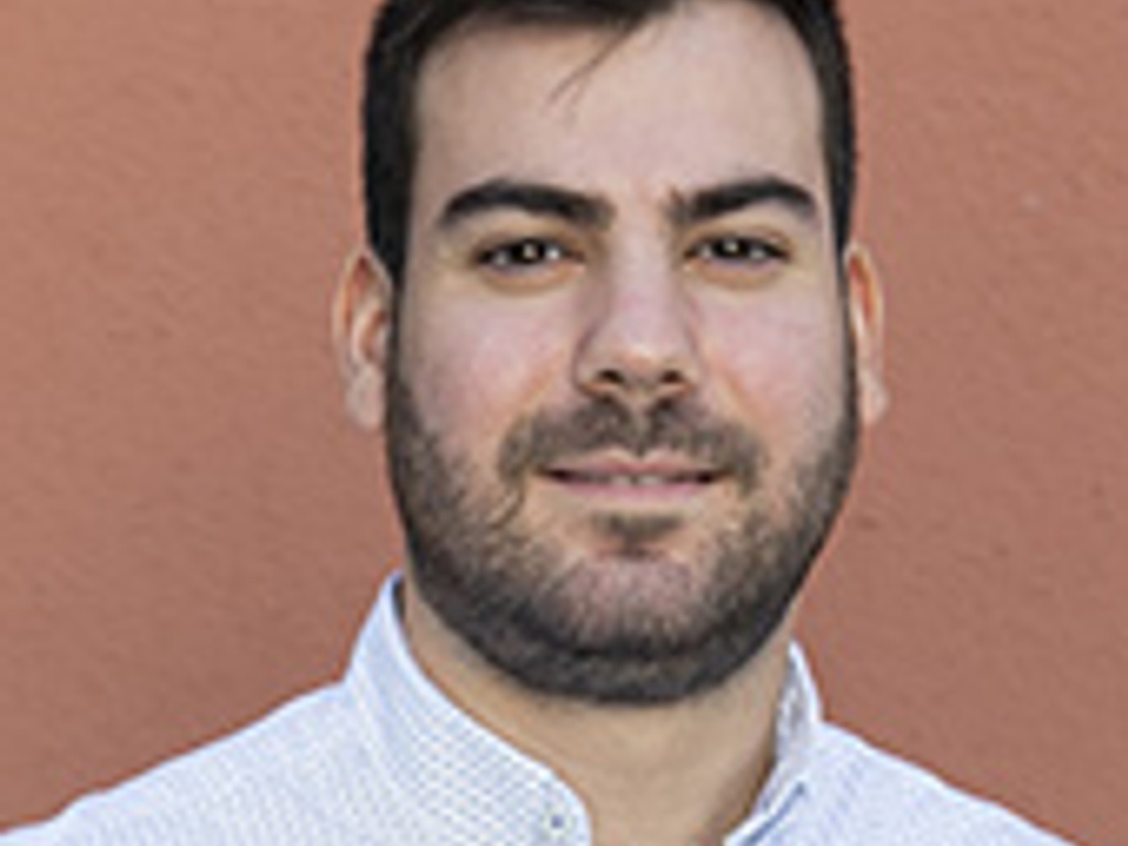 A close-up portrait of a smiling man with a beard wearing a white dotted shirt against a pink background.