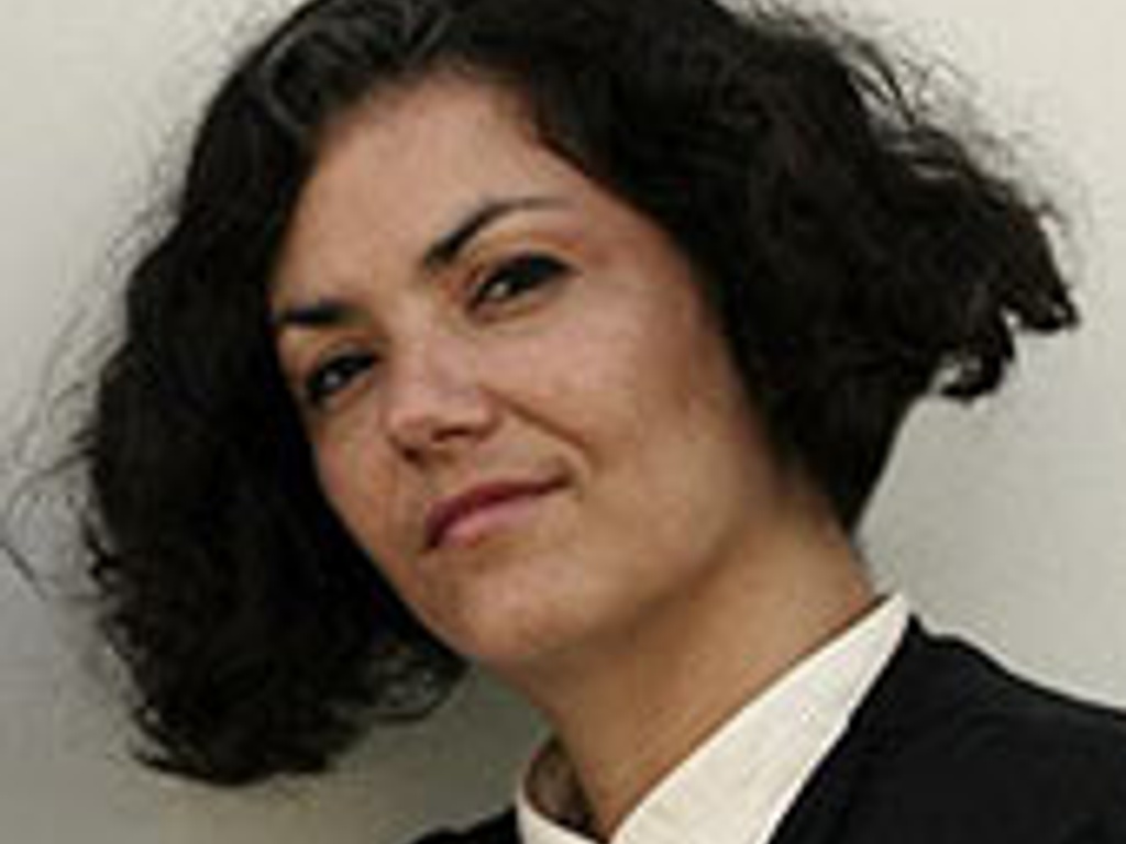 A portrait of a confident woman with curly hair, wearing a white shirt and black blazer, looking directly at the camera.